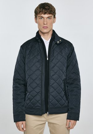 Gequilted donkerblauwe jas met een ritssluiting aan de voorkant, opstaande kraag en gouden drukknoopjes. Voorzien van zijzakken en geribbelde manchetten.