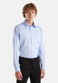 Camicia azzurro chiaro a maniche lunghe con colletto alla francese, realizzata in un tessuto liscio; indossata con jeans neri, dotati di tasche frontali.