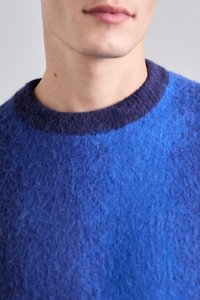 Zila džemperis ar teksturētu adījumu, kas pāriet no tumši zila uz gaišākām nokrāsām apkaklītē. Apaļa kakla izgriezums, maiga apdare.