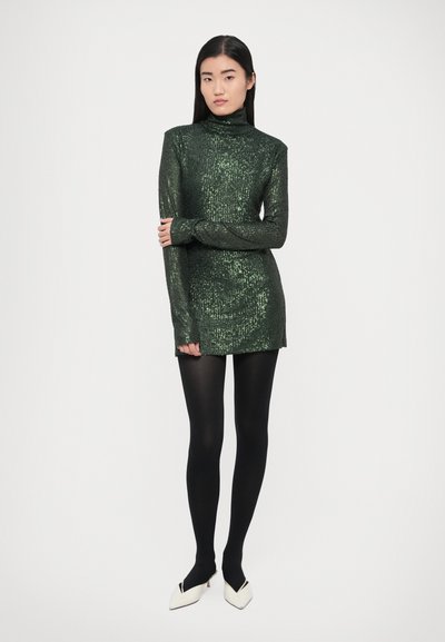 Zöld, hosszú ujjú ruha magas nyakkal és flitteres textúrával, fekete harisnyával és fehér hegyes orrú magassarkúval párosítva.