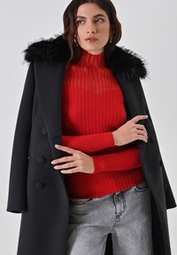 Schwarzer Wollmantel mit einem Kragen aus Kunstpelz, über einem leuchtend roten gerippten Pullover getragen. Hellgraue Jeans vervollständigen das Outfit.