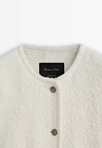 Cardigan en tricot blanc texturé avec encolure ronde et deux boutons métalliques décoratifs à l'avant, étiquette à l'intérieur du col.