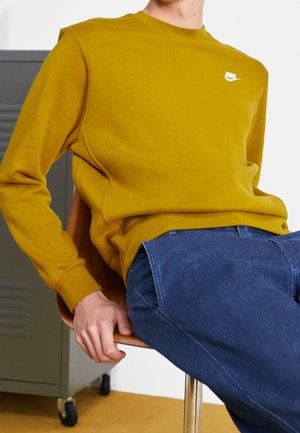 Mosterdgele sweatshirt met een ronde hals en wit logo, gecombineerd met donkerblauwe textuur jeans, zittend op een houten stoel.