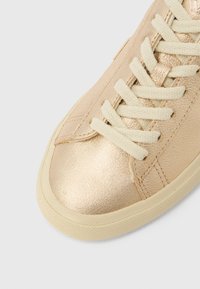 Foran nærbilde av en gullfarget metallisk sneaker med kremhvite lisser og beige gummisåle på hvit bakgrunn.