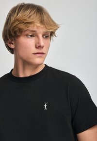 Czarna bawełniana koszulka z okrągłym dekoltem, z małym białym logo golfisty na lewej piersi, gładka tekstura i krótkie rękawy.