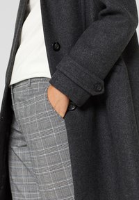 Abrigo de lana gris con botones dobles, con un acabado texturizado. Combinado con pantalones a cuadros en gris y negro, con la mano en el bolsillo.