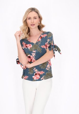 Blomsterprint v-hals top i teal med pink og grønne mønstre. Har korte ærmer med bindebånd. Båret sammen med hvide bukser, glat stofstruktur.