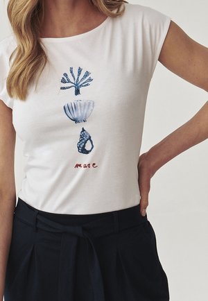 Frau trägt ein weißes Kurzarmoberteil mit blauem Korallen- und Muscheldruck sowie rotem Text, kombiniert mit hoch taillierten schwarzen Hosen mit Gürtel-Detail.