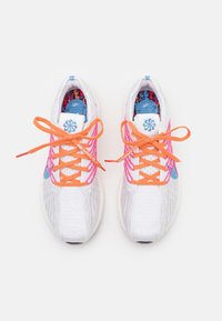 Baskets tricotées blanches avec des détails roses, lacets orange vif noués en nœuds, et semelles intérieures colorées bleues et rouges, vues de dessus.