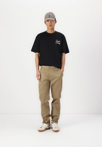 Črna bombažna majica s belim napisom, bež chino hlače in bele superge z rjavimi detajli. Model nosi sivo kapo. Preprost, sproščen outfit.