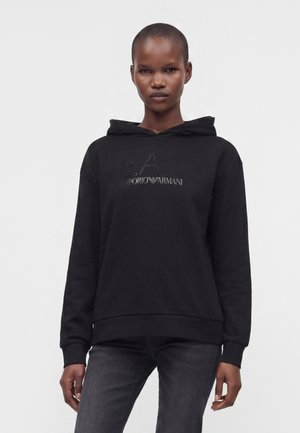 Femme aux cheveux courts portant un sweat à capuche noir Emporio Armani EA7 et un jean sombre, debout devant un fond clair uni.