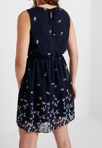 Námořnická modrá bezramenní šaty s potiskem motýlů, elastickým pasem a detailem na zavazování vzadu; má volnou, průhlednou sukni.