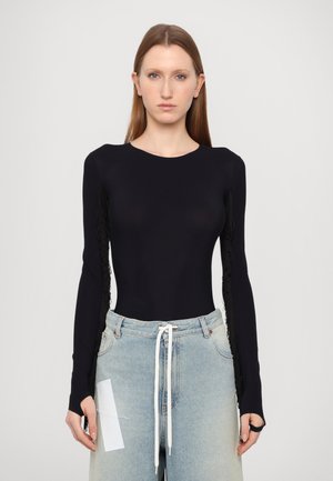 Bodysuit noir à manches longues avec des franges sur les manches, associé à un jean taille haute bleu clair et un cordon blanc. Texture lisse.