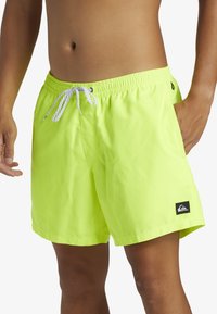 Bañador verde neón con cintura elástica, cordón ajustable, bolsillos laterales y un parche de logo negro en la pierna izquierda.