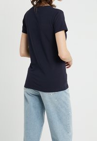 Kobieta ubrana w granatową koszulę z krótkim rękawem i jasnoniebieskie dżinsy, stojąca z odwróconymi plecami i zgiętymi ramionami.