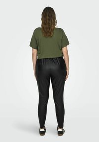 Olijfgroene top met korte mouwen, gecombineerd met zwarte, strakke neplederen broek. Opvallende gladde textuur en taps toelopend ontwerp van de pijp. Casual schoenen zichtbaar.