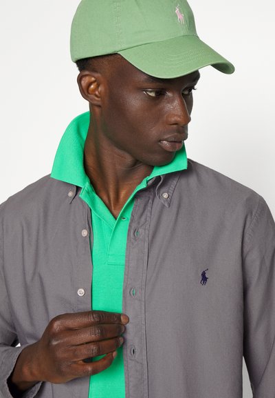 Camisa gris de botones sobre un polo verde, con una gorra verde. Presenta bordado de logotipo tanto en la camisa como en la gorra. Texturas suaves.