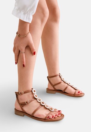 Jambes portant des sandales plates marron ornées de perles circulaires décoratives, main reposant sur le mollet, vernis à ongles rouge sur les doigts et les orteils.