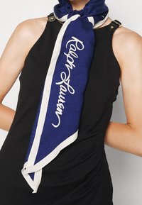 Námořnický modrý hedvábný šátek s bílými okraji, na kterém je logo "Ralph Lauren" napsané kurzívou. Uvázaný kolem krku, v kontrastu s černým topem.