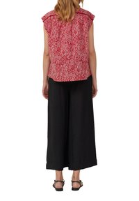 Rode blouse met patroon en korte mouwen, voorzien van een plooi aan de achterkant, gecombineerd met een zwarte wijde broek en platte sandalen.