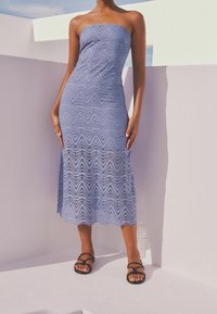 Robe sans bretelles mi-longue de couleur bleu clair, ornée d'un motif en chevron avec des découpes. Portée avec des sandales noires à lanières sur fond blanc.