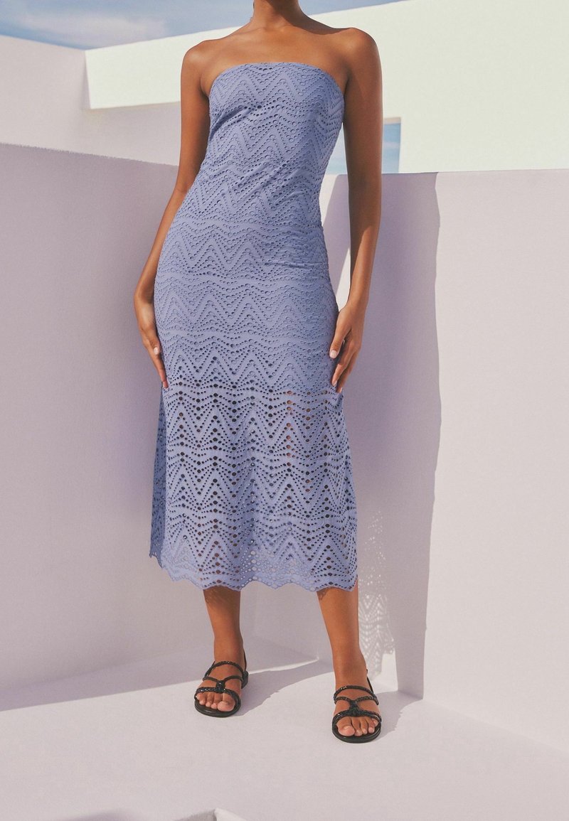 Robe sans bretelles mi-longue de couleur bleu clair, ornée d'un motif en chevron avec des découpes. Portée avec des sandales noires à lanières sur fond blanc.