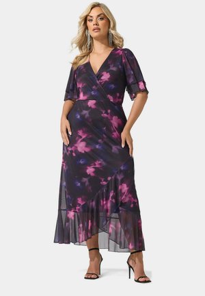Mujer con cabello rubio que lleva un vestido cruzado floral en tonos morado y rosa con volante en el dobladillo y sandalias negras de tacón, de pie frente a un fondo liso.