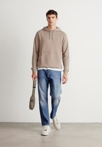 Jonge man loopt binnenshuis en draagt een beige hoodie, blauwe jeans, witte sneakers en houdt een bijpassende beige schoudertas vast.