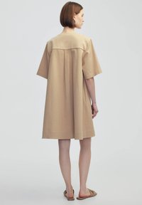 Vestido beige de manga corta con tejido texturizado, silueta suelta y una costura en los hombros. La modelo lleva sandalias color tostado.