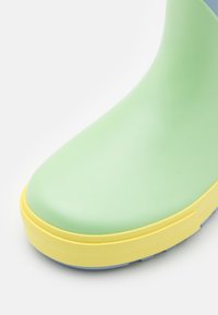 Grüner Gummistiefel mit runden Zehen, ausgestattet mit einer gelben Außensohle und blauen oberen Akzenten. Glatte Textur, minimalistisches Design und keine sichtbaren Muster.