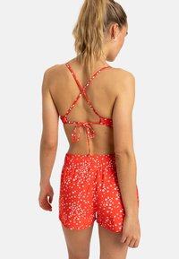 Röd bikinitäcke med korsade band, knytet i ryggen; matchas med röda shorts med ett vitt blommönster och elastisk midja.