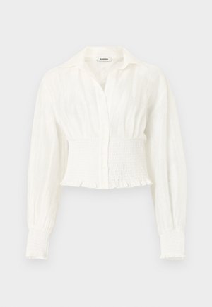 Blouse blanche à manches longues avec taille et poignets froncés, devant boutonné et col pointu sur un fond uni.