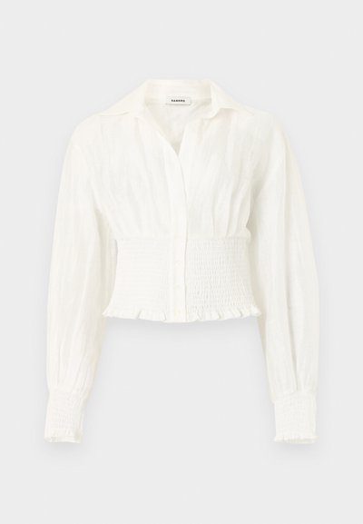 Blouse blanche à manches longues avec taille et poignets froncés, devant boutonné et col pointu sur un fond uni.