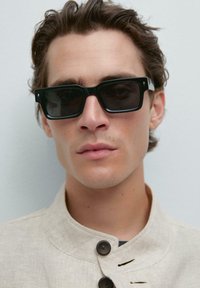 Occhiali da sole neri rettangolari con lenti scure, caratterizzati da dettagli bianchi sulle tempie. Il modello indossa una giacca beige con colletto.