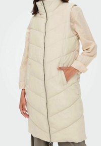 Długi beżowy kamizelka puchowa z wysokim kołnierzem, z centralnym zamkiem, wzorem w zygzak oraz bocznymi kieszeniami, wykonana z gładkiego, pikowanego materiału.
