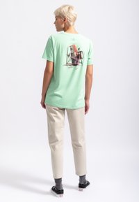 T-shirt en coton vert menthe avec un imprimé graphique au dos. Associé à un pantalon beige et des chaussures noires, mettant en avant une coupe et un design décontractés.