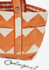 Narancssárga és fehér cikkcakk mintázatú vászontáska, szőtt textúrával, díszítéssel és fogantyúkkal. Ezüst színű kulcstartó részletet tartalmaz.