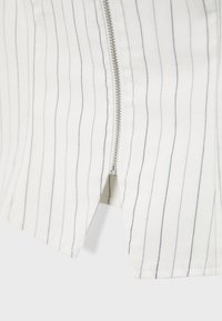 Fermeture éclair métallique partiellement ouverte sur un tissu blanc avec de fines rayures verticales noires.