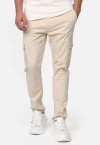 Pantalons cargo beiges en tissu léger avec poches latérales et coupe fuselée. Portés avec des chaussures de sport blanches présentant des accents en maille.