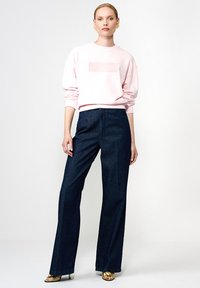 Hele roosa sweatshirt, millel on reljeefne logo, koos tumeda sinise laia jala teksapükstega. Mudel kannab kuldseid kontsaga sandaale. Lihtne taust.