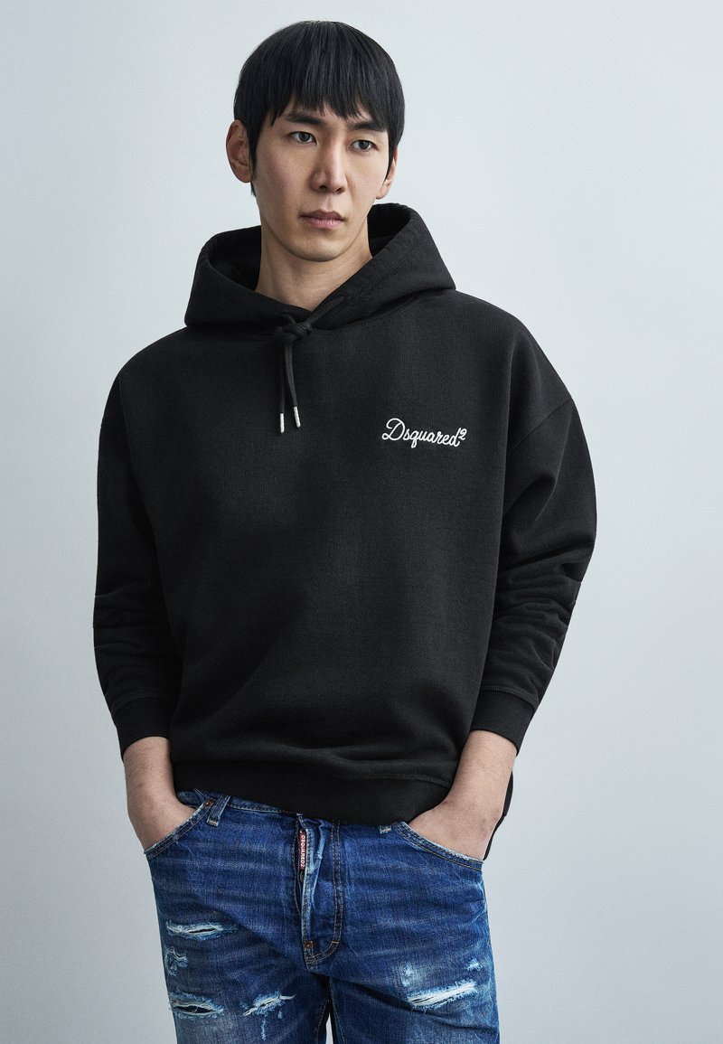 Μαύρο φούτερ με κορδόνι που διαθέτ ει λευκό λογότυπο «Dsquared2» στο στήθος. Φορεμένο με φθαρμένα μπλε τζιν. Ομαλή υφή υφάσματος.