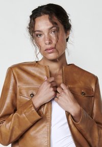 Giubbotto in pelle marrone con collo tondo, chiusura con zip e due tasche frontali. Texture liscia e design minimalista.