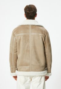 Chaqueta de ante beige con un cuello y puños de piel de oveja blanca. Presenta una textura suave, tirantes ajustables en el dobladillo y mínimos detalles de costura.