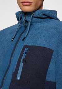 Flisani zip-up dukserica u nijansama plave boje s teksturiranim završetkom, s tamnoplavim detaljem i podesivim kapuljačom.