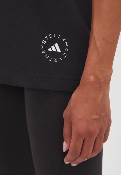 T-shirt de sport noir avec un logo circulaire blanc comportant du texte et trois rayures sur l'ourlet, porté avec des leggings noirs.
