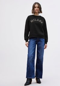 Sudadera negra con el texto "MILANO" en color claro, combinada con vaqueros anchos de mezclilla azul y botines negros. Textura suave, diseño casual.