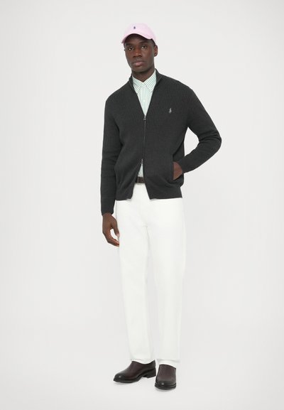 Sötétszürke bordázott kardigán, cipzáras záródással, világoszöld csíkos ing fölött viselve, fehér nadrággal, barna csizmával és rózsaszín sapkával.