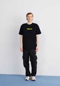 Čierne tričko s neónovo zeleným logom "ellesse", spárované s čiernymi cargo nohavicami s bočnými vreckami a žltými akcentmi. Bielene tenisky.