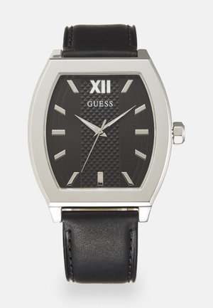 Ceas rectangular negru cu accente din metal argintiu, față texturată de culoare închisă, marcaje albe pentru ore și curea din piele neagră. Logo-ul brandului: Guess.