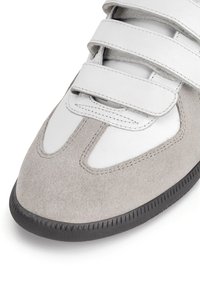 Hall hall ja valge nahkste sneaker, millel on kolm Velcro rihma, ümar nina ja tekstureeritud kummist põhi haardeks.